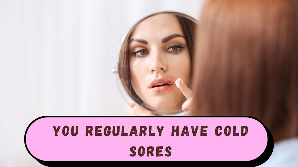 Girl looking at themselves in a mirror with a worried expression, showing a visible cold sore on their lip, with a small tube of ointment on the counter.
